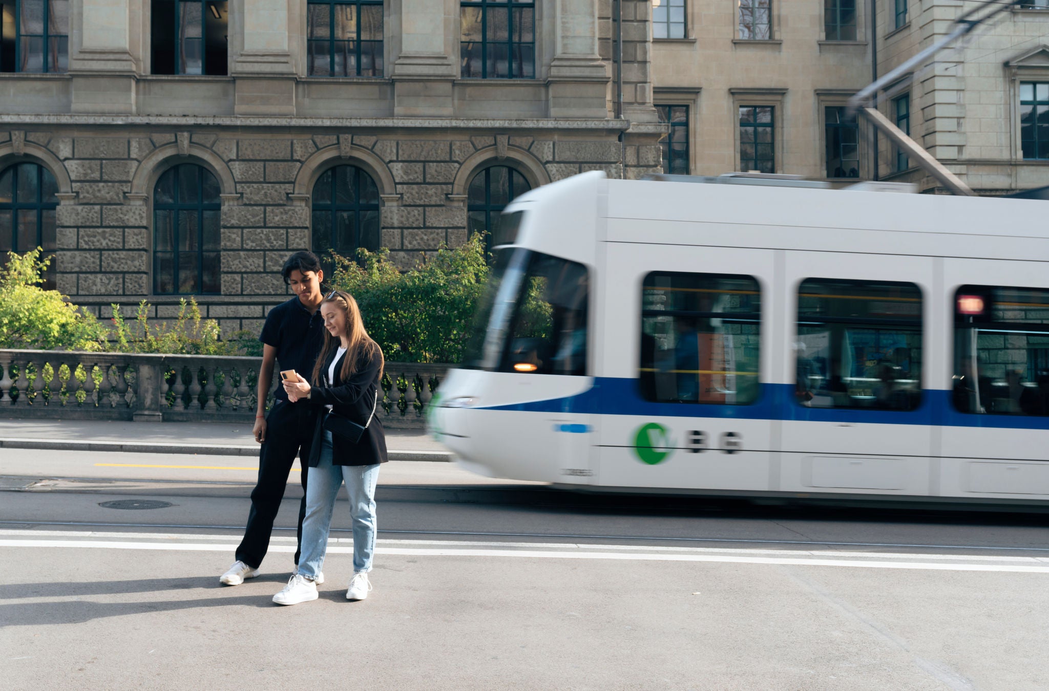 Ein junges Paar steht an der Tramhaltestelle und schaut aufs Smartphone.